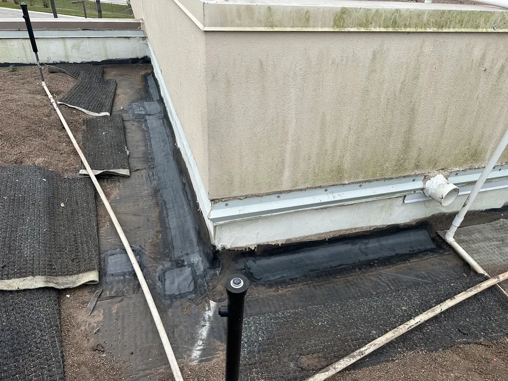 Completed base flashing repair where a low-slope commercial roof meets a stucco parapet, black reinforced membrane run along the base of the wall and around the corner, with walkway pads and pipes on the roof